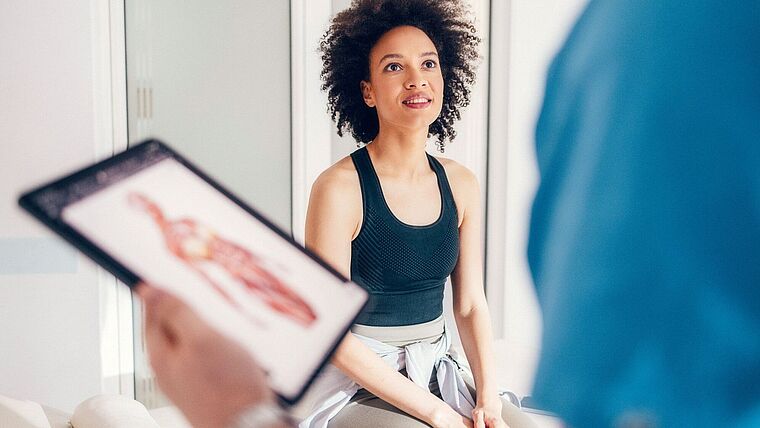 Physio hät tablet mit Bodychart und schaut eine Patientin dabei an, die in die Kamera blickt