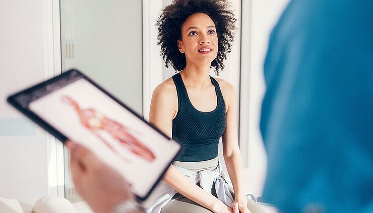 Physio hät tablet mit Bodychart und schaut eine Patientin dabei an, die in die Kamera blickt